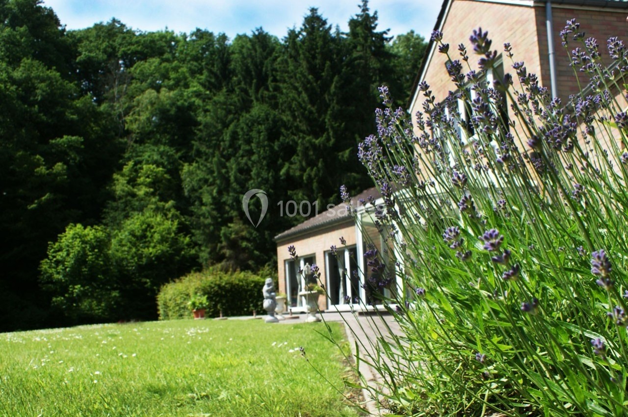 Jardin verdoyant avec lavandes au premier plan, maison en briques et forêt en arrière-plan sous un ciel dégagé.