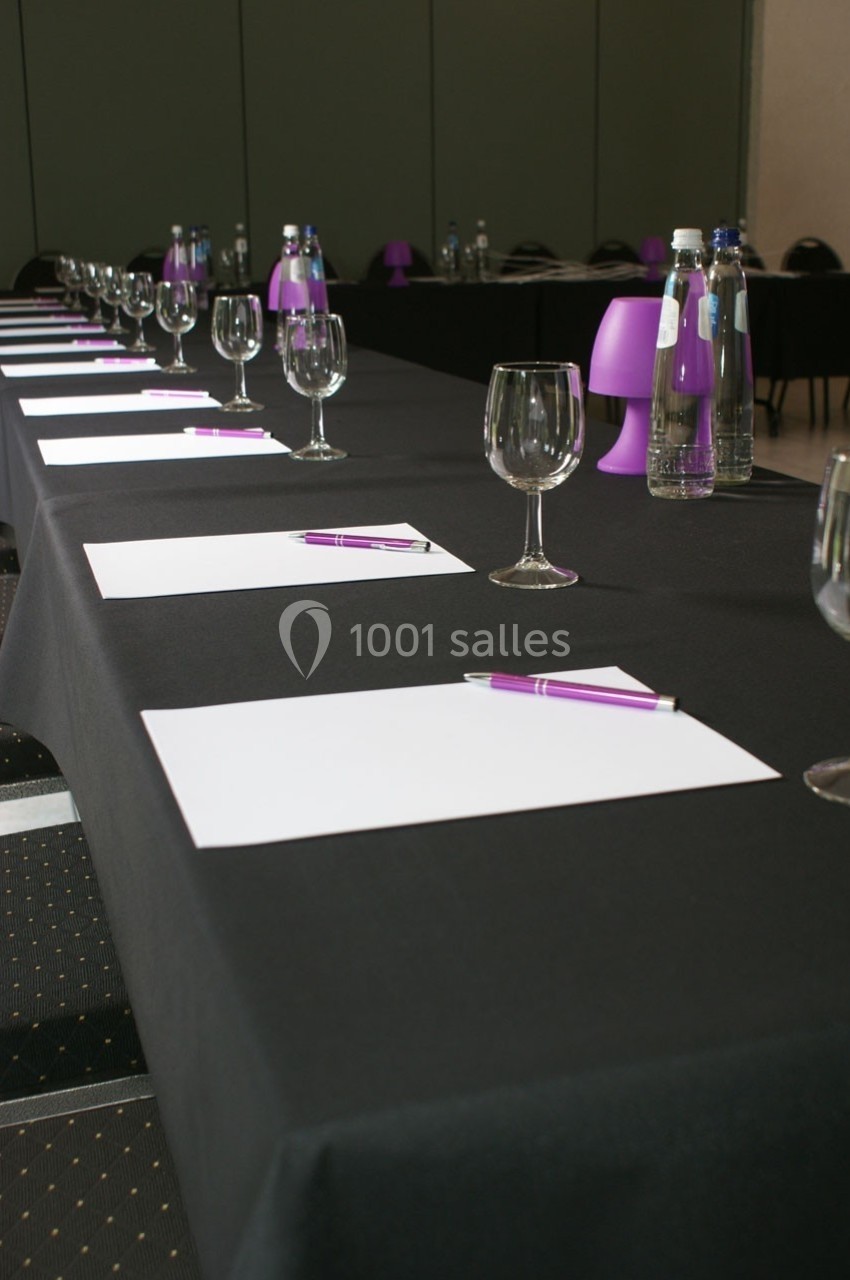 Salle de réunion avec une table noire dressée de verres, bouteilles d'eau, feuilles blanches et stylos violets.