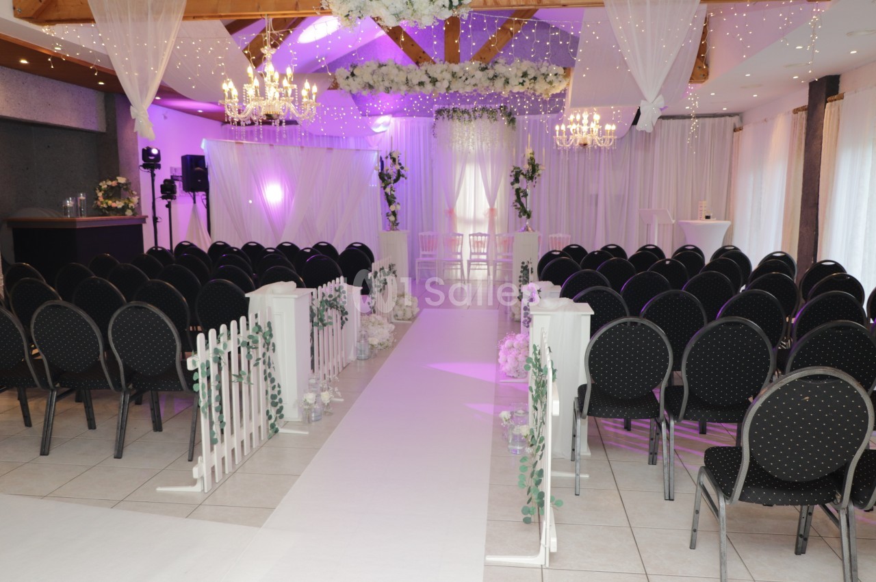 Salle de réception dressée pour une cérémonie avec rangées de chaises noires de part et d'autre d'une allée blanche, arche fleurie et voilages sous un éclairage violet.
