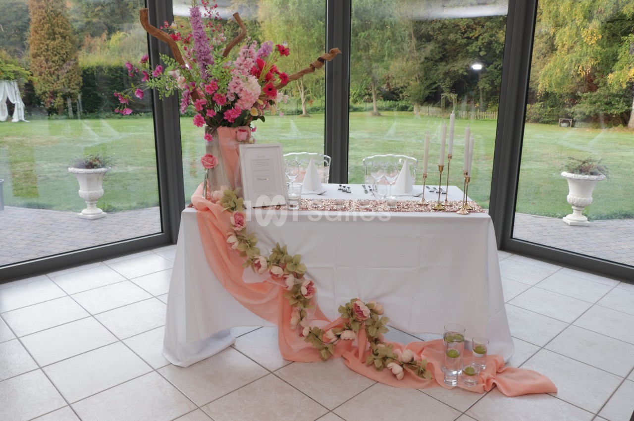 Table de réception nappée de blanc et de sequins rose, décorée de fleurs roses et d'un voilage saumon, devant de grandes baies vitrées donnant sur un jardin.