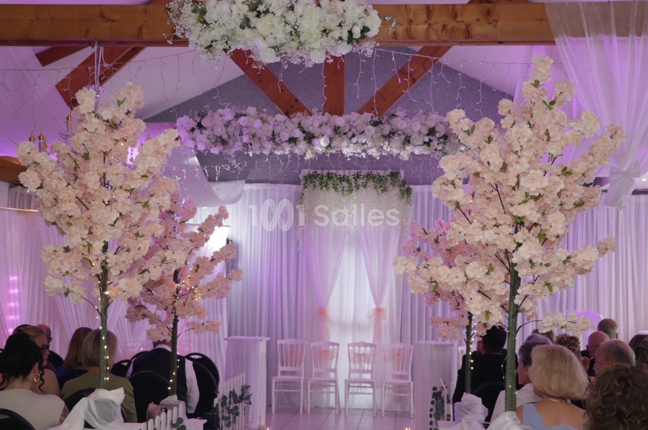 Salle de réception décorée de grands arbres artificiels en fleurs blanches et roses, avec une arche fleurie, des rideaux blancs et des chaises face à une estrade éclairée en violet.