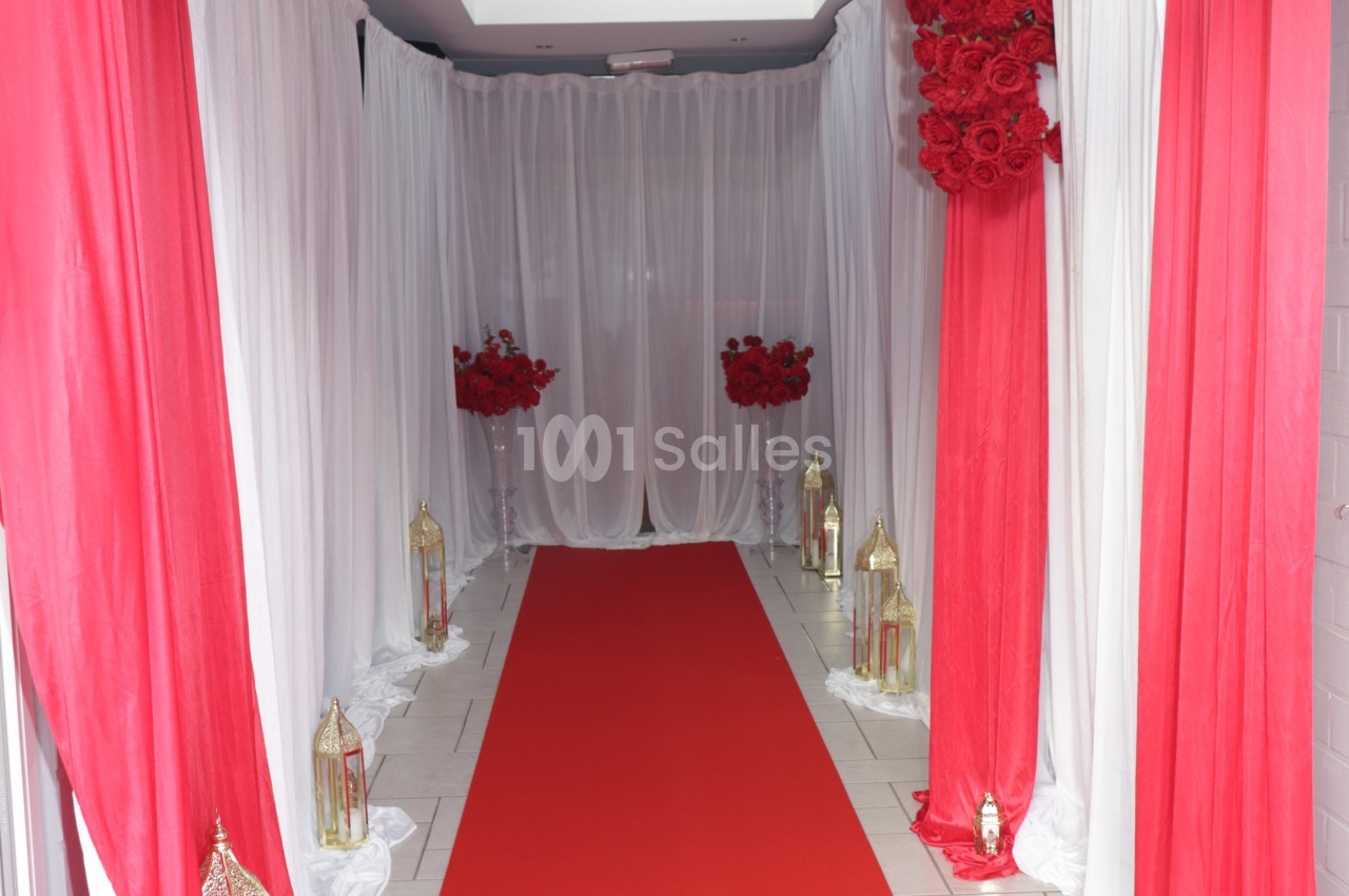 Allée d'entrée décorée de voilages blancs et rouges, avec tapis rouge, roses rouges en hauteur et lanternes dorées au sol.