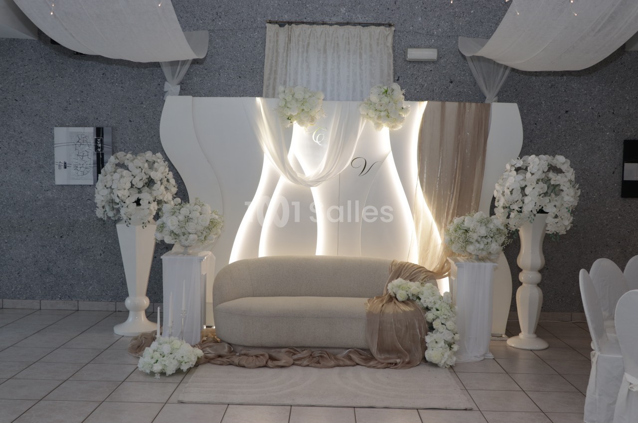 Scène de mariage aménagée : un canapé beige devant un panneau blanc à éclairage LED, orné de hortensias blancs sur des vases et colonnes, et de voilages drapés. Let me check: under 125 characters? Count roughly: "Scène de mariage aménagée : un canapé