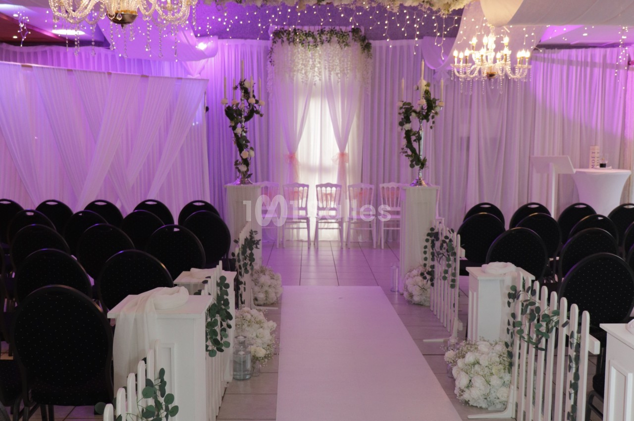 Salle de réception décorée de blanc et de mauve pour une cérémonie, avec allée centrale tapissée, rangées de chaises noires, bordures fleuries et lustres en cristal.