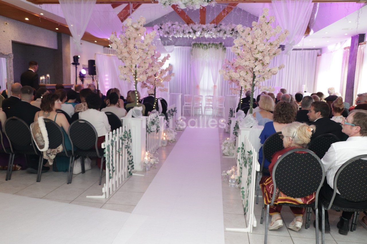 Salle de cérémonie avec allée blanche centrale, arches de fleurs blanches et rangées de chaises tournées vers l'autel, sous un éclairage violet.