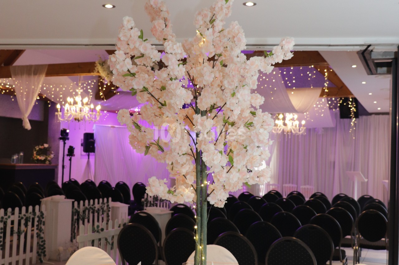 Grand arbre artificiel à fleurs blanches et roses avec guirlandes lumineuses, au centre d'une salle de réception avec chaises noires rembourrées en rangs.