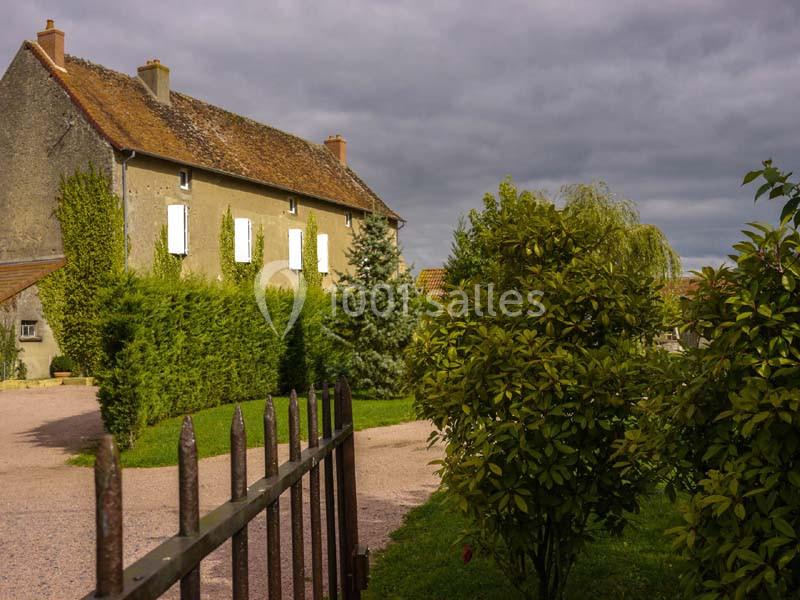 Maison en pierre avec volets blancs entourée de haies, arbres et buissons, sous un ciel nuageux.