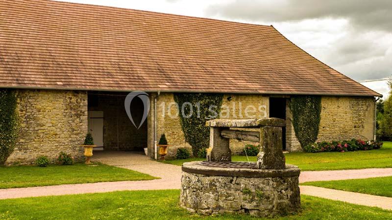 Une ancienne grange en pierre avec un puits en pierre au premier plan, entourée de pelouse et de végétation.