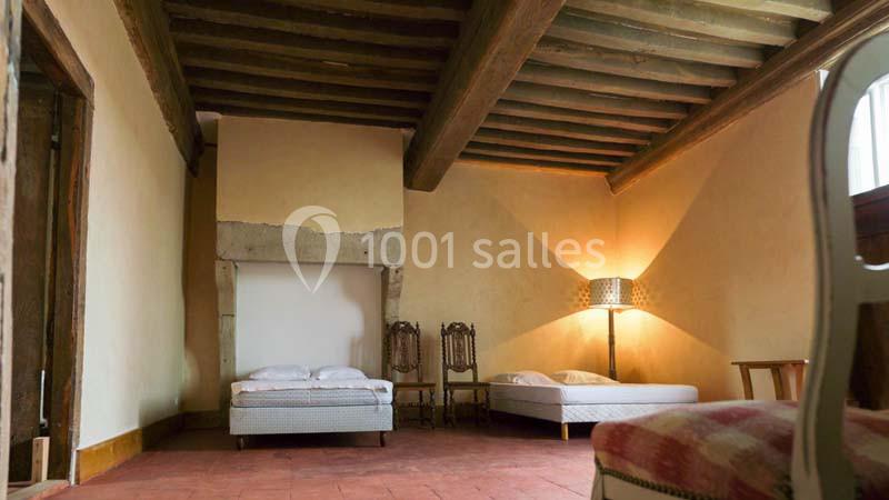 Chambre avec deux lits simples, poutres apparentes, sol en terre cuite et lampe allumée près d'un mur beige.