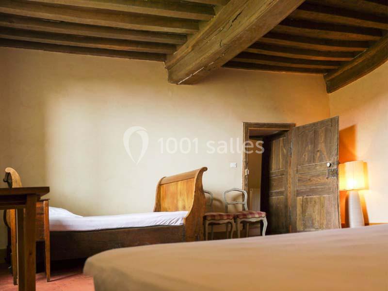 Chambre rustique avec lits en bois, plafond à poutres apparentes, chaises anciennes et lampe sur pied.