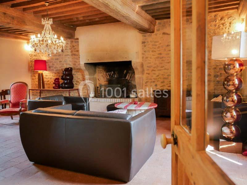 Salon chaleureux avec murs en pierre, cheminée, lustre, fauteuils rouges et canapés en cuir sombre.