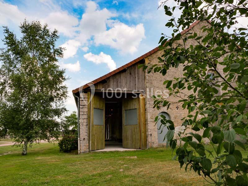 Grange en pierre avec portes ouvertes, entourée d'arbres et d'herbe sous un ciel partiellement nuageux.