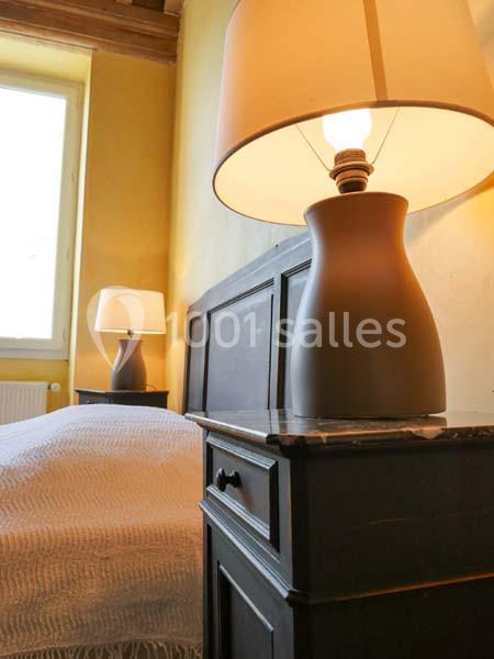 Lampe de chevet allumée sur une table en bois sombre près d'un lit, dans une chambre à la lumière naturelle.