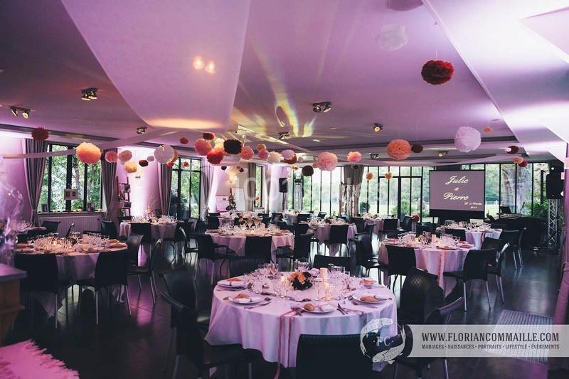 Salle de réception décorée avec des tables rondes, nappes blanches et guirlandes de pompons suspendues au plafond.
