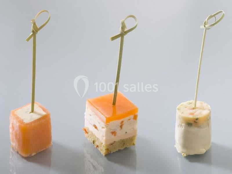 Trois amuse-bouches sur piques en bois, composés de saumon, gelée orange et préparation crémeuse, sur fond neutre.