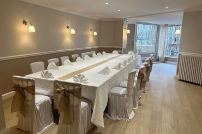 Salle lumineuse avec une grande table rectangulaire dressée, entourée de chaises décorées, parquet clair et fenêtres donnant…