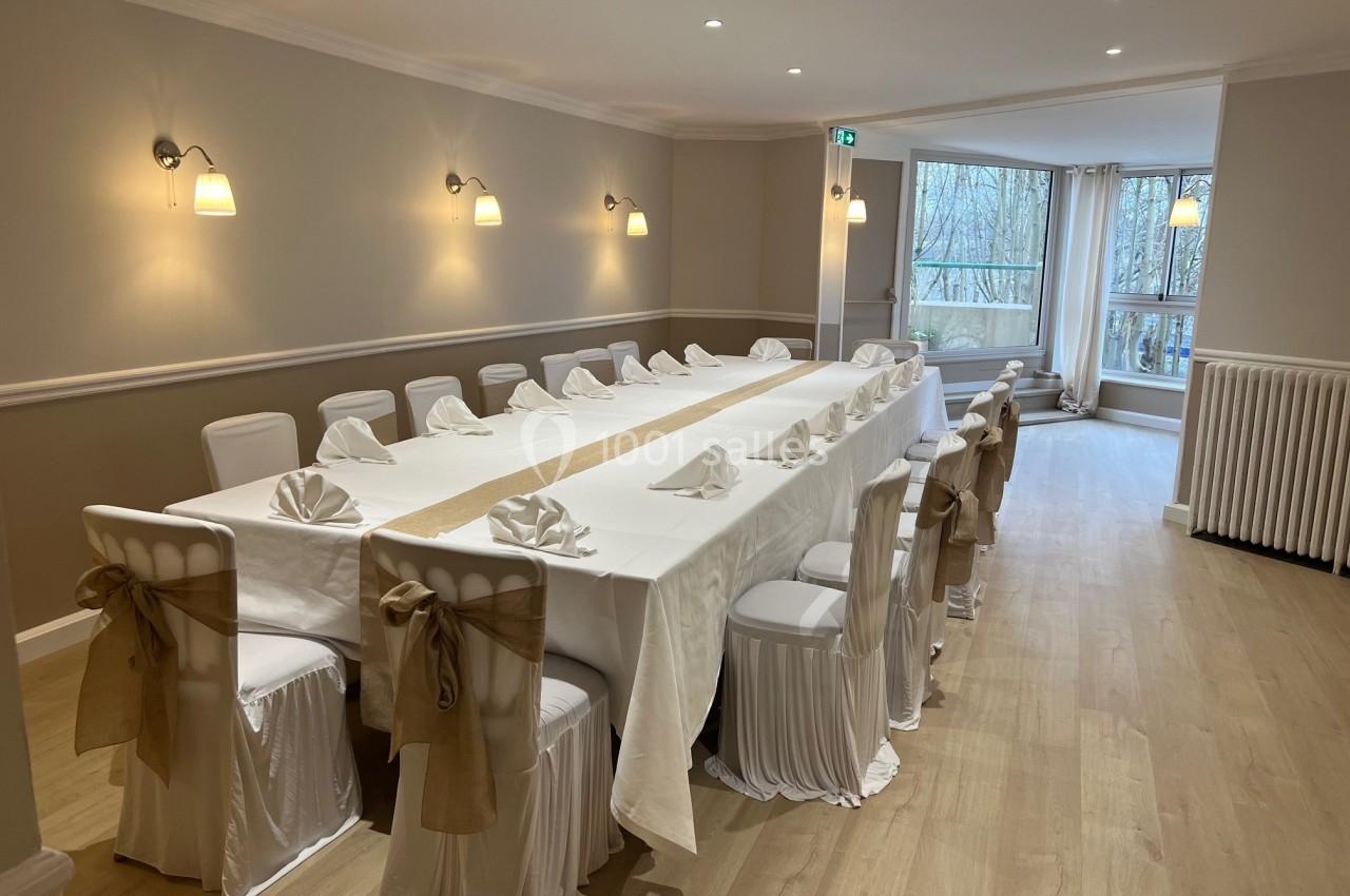 Salle lumineuse avec grande table rectangulaire dressée, chaises blanches décorées de nœuds en tissu beige.