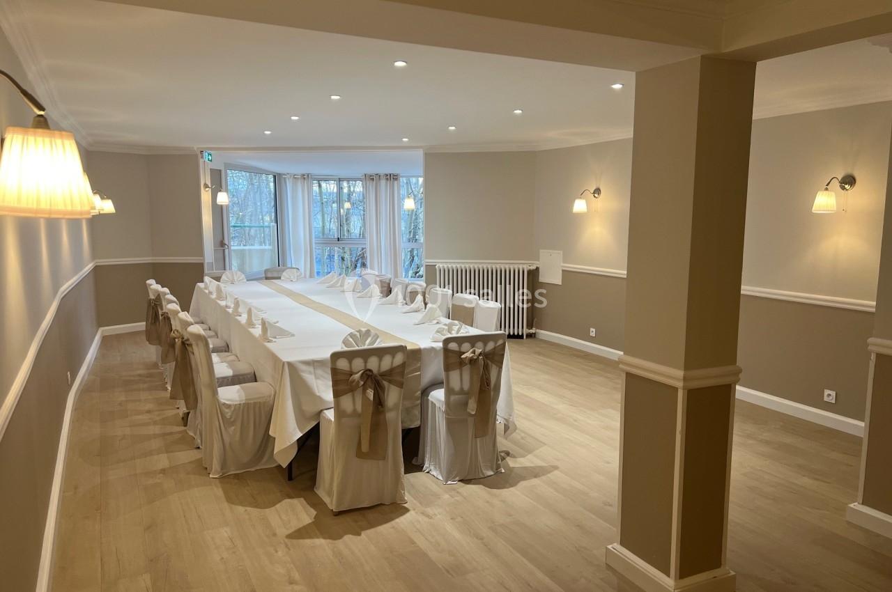 Salle lumineuse avec une grande table rectangulaire dressée, entourée de chaises décorées, parquet clair et fenêtres donnant…
