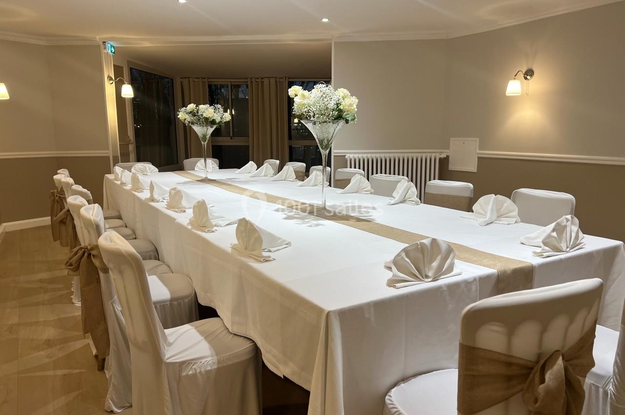 Salle de réception avec une grande table dressée, nappes blanches, serviettes pliées et décorations florales.