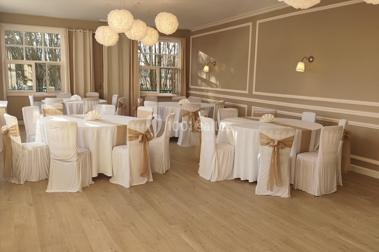 Salle de réception lumineuse avec tables rondes dressées, chaises blanches ornées de nœuds dorés et suspensions décoratives.