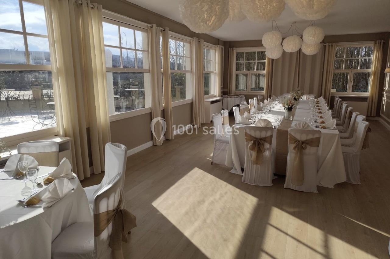 Salle lumineuse avec de grandes fenêtres, tables dressées pour un repas, chaises décorées de nœuds dorés.