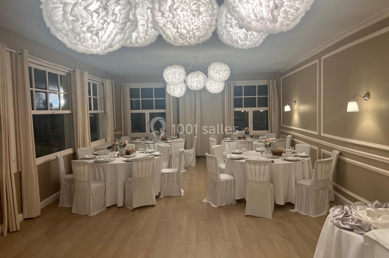 Salle de réception élégante avec tables rondes dressées, chaises blanches et suspensions lumineuses décoratives.