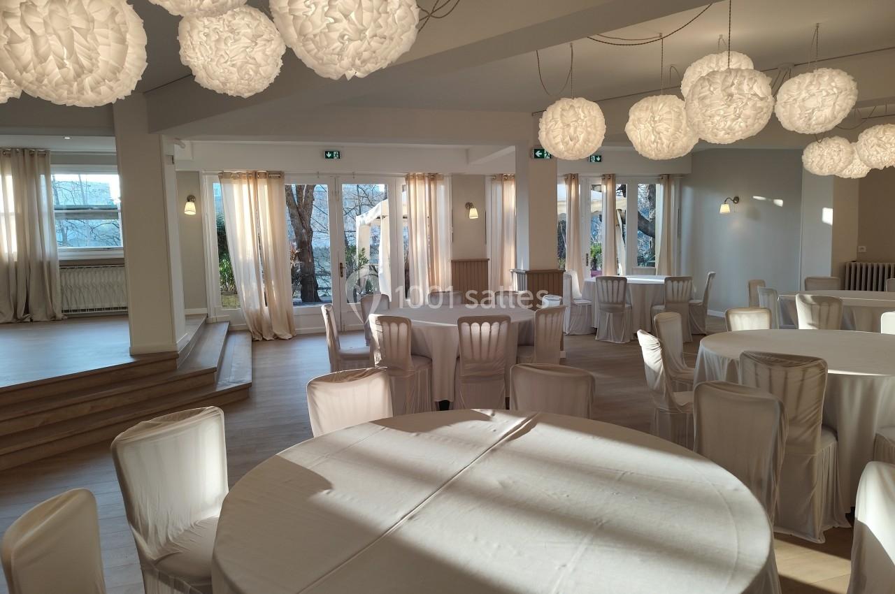 Salle de réception lumineuse avec tables rondes dressées de nappes blanches et suspensions décoratives.