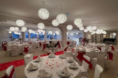Salle lumineuse avec une grande table rectangulaire dressée, entourée de chaises décorées, parquet clair et fenêtres donnant…