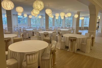 Salle lumineuse avec une grande table rectangulaire dressée, entourée de chaises décorées, parquet clair et fenêtres donnant…