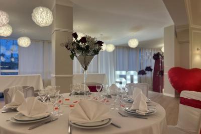 Salle lumineuse avec une grande table rectangulaire dressée, entourée de chaises décorées, parquet clair et fenêtres donnant…