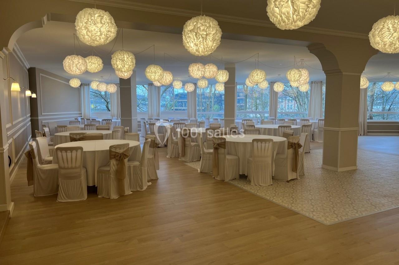 Salle de réception lumineuse avec tables rondes dressées et chaises blanches décorées de nœuds beiges.