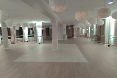 Salle lumineuse avec une grande table rectangulaire dressée, entourée de chaises décorées, parquet clair et fenêtres donnant…