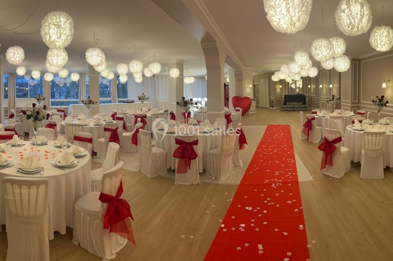 Salle de réception décorée avec des tables rondes, nappes blanches, nœuds rouges et un tapis rouge au centre.
