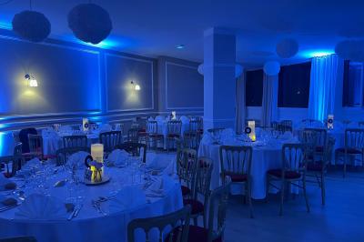 Salle lumineuse avec une grande table rectangulaire dressée, entourée de chaises décorées, parquet clair et fenêtres donnant…