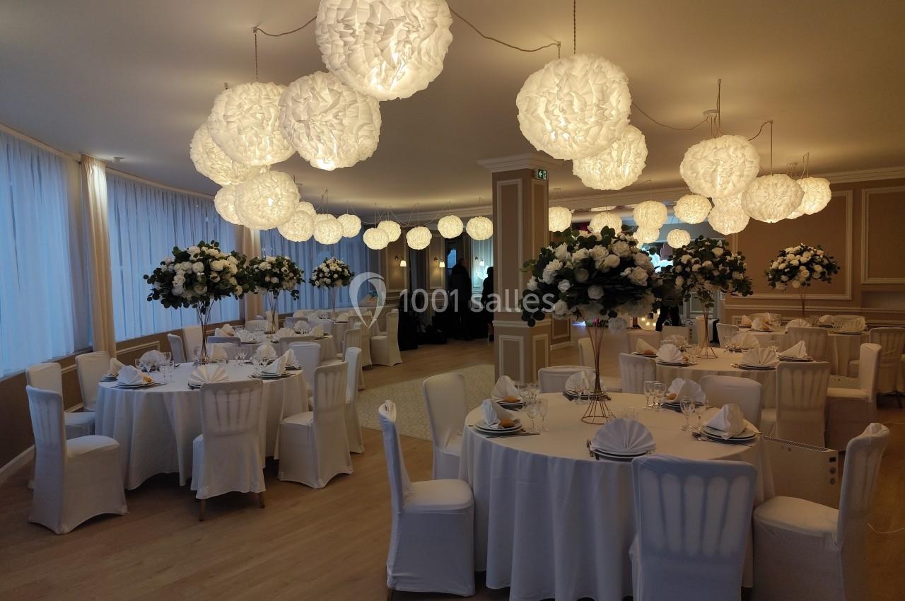 Salle de réception élégante avec tables rondes dressées, chaises blanches et décorations florales sous des luminaires…