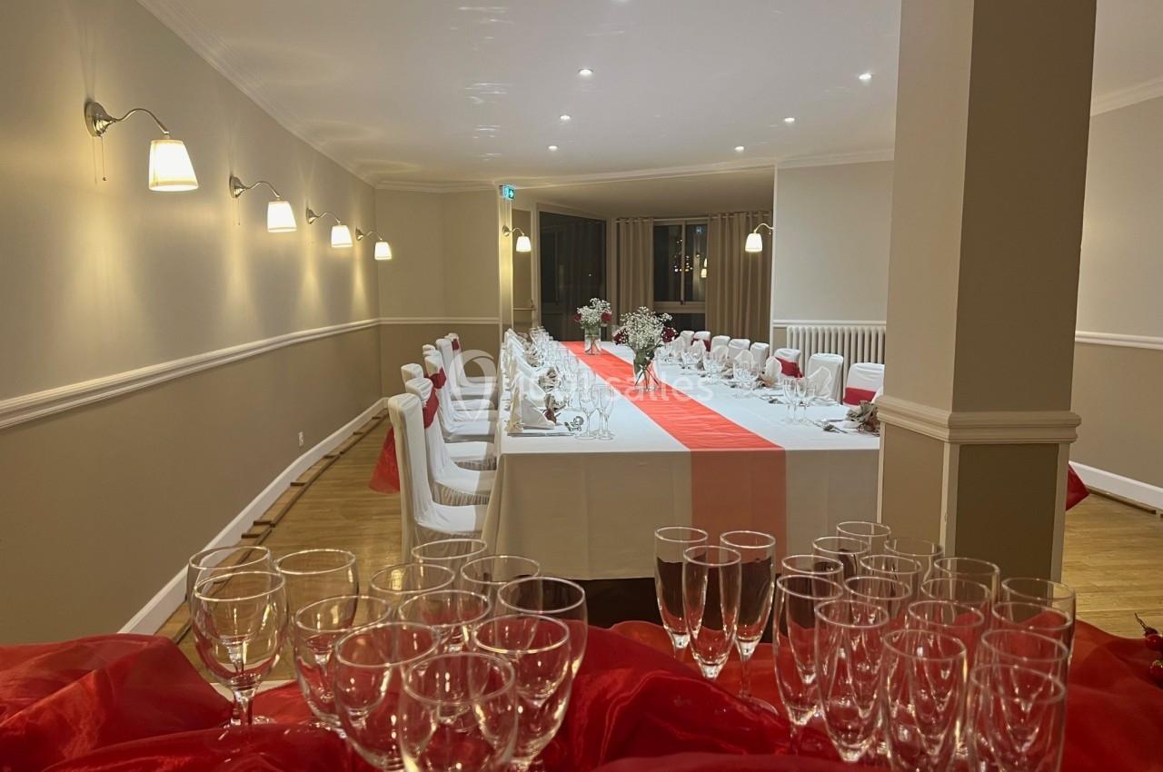 Salle de réception décorée avec une longue table dressée, nappe blanche et chemin de table rouge, verres en premier plan.