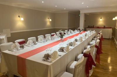 Salle lumineuse avec une grande table rectangulaire dressée, entourée de chaises décorées, parquet clair et fenêtres donnant…