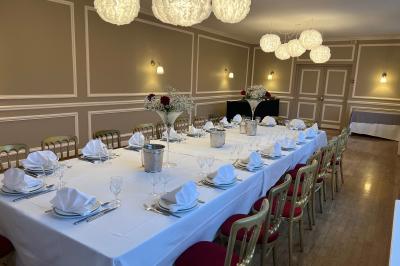 Salle lumineuse avec une grande table rectangulaire dressée, entourée de chaises décorées, parquet clair et fenêtres donnant…