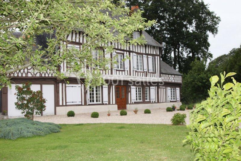 Façade d'une maison à colombages entourée d'un jardin avec pelouse, arbres et allée gravillonnée.