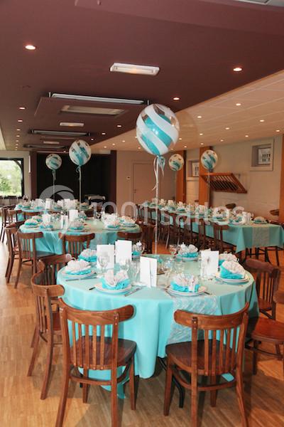 Salle décorée pour un événement avec des tables rondes dressées en bleu et blanc, ornées de ballons assortis.