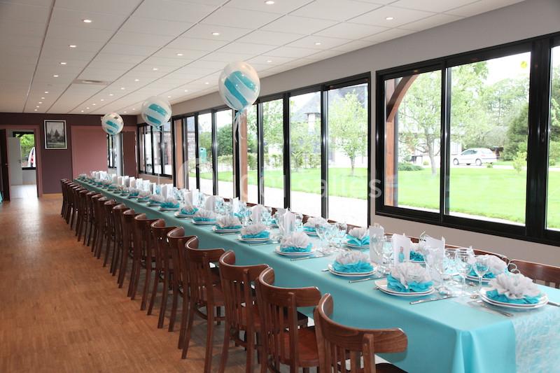 Salle décorée pour un événement avec une longue table dressée, nappes bleues, serviettes pliées et ballons assortis.