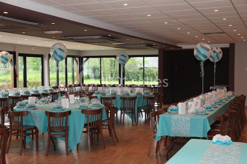 Salle décorée pour un événement avec des tables dressées en bleu et blanc, ballons suspendus et grandes baies vitrées.
