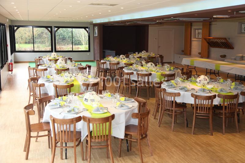 Salle de réception avec des tables rondes dressées, nappes blanches et chemins de table verts, éclairée par des baies…