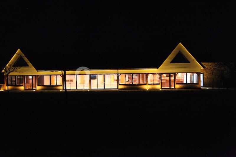 Bâtiment illuminé de nuit avec des façades vitrées et un toit en pente, entouré d'obscurité.