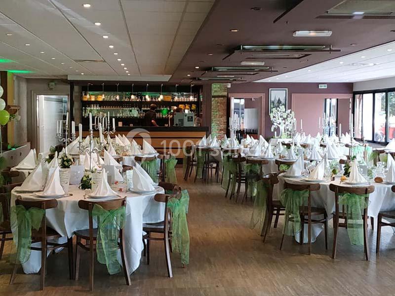 Salle de réception décorée avec des tables rondes, nappes blanches, serviettes pliées et chaises ornées de rubans verts.