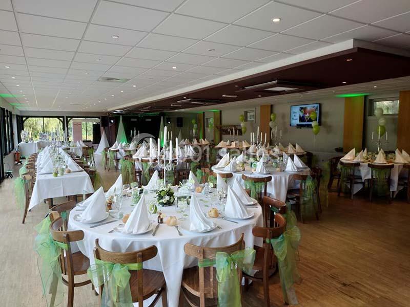 Salle de réception décorée avec des tables rondes, nappes blanches, serviettes pliées et chaises ornées de rubans verts.