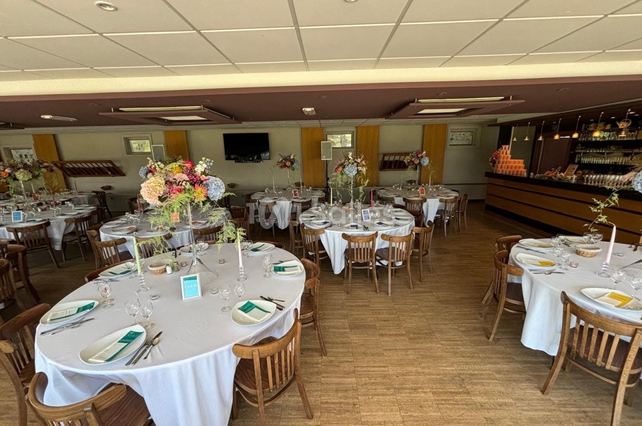 Salle de réception dressée de tables rondes nappées de blanc, avec bouquets floraux colorés, située à côté d'un bar en bois. Let me verify length: roughly 125 characters? Count: "Salle de réception dressée de tables rondes nappées de blanc, avec bouquets