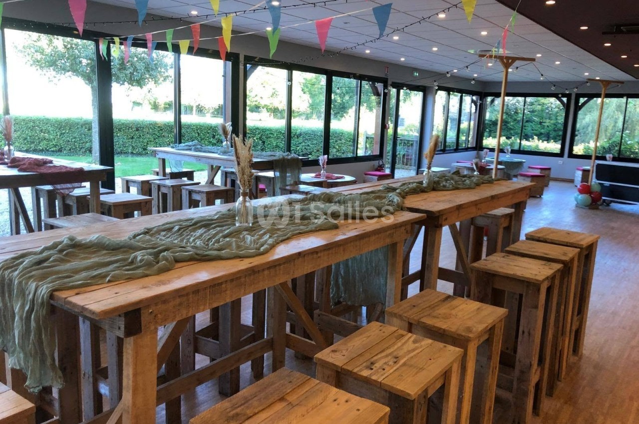 Salle lumineuse aux grandes baies vitrées, dressée de longues tables hautes en bois brut avec tabourets, chemins de table gaze vert sauge et guirlandes de fanions.