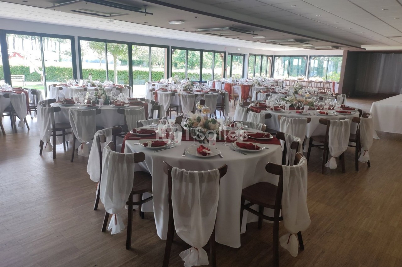 Salle de réception lumineuse avec de nombreuses tables rondes dressées de nappes blanches, serviettes rouges et chaises habillées de voiles blancs, vue sur un parc par de grandes baies vitrées.