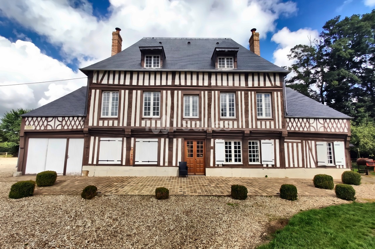 Façade à colombages blanc et brun d'une grande demeure normande, avec volets blancs, deux cheminées et un pavage devant l'entrée.
